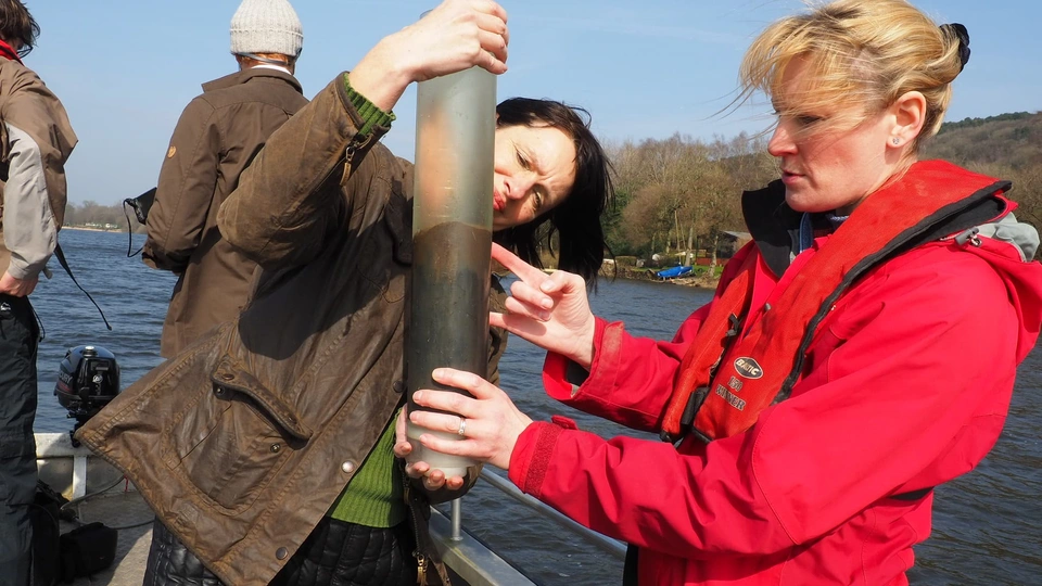 Two female academics looking into a tube with sediment sample inside whilst on a boat