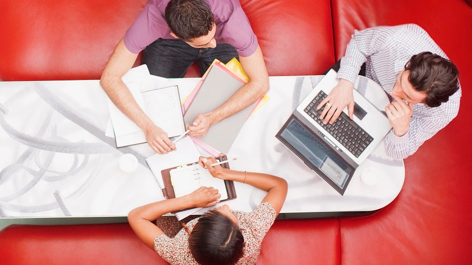 A group of three people are sat on a red sofa around a desk, using their laptops