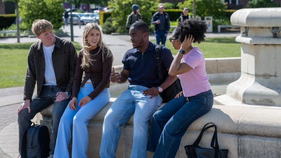 A group of four students are sat on the edge of the University fountain, smiling and chatting together