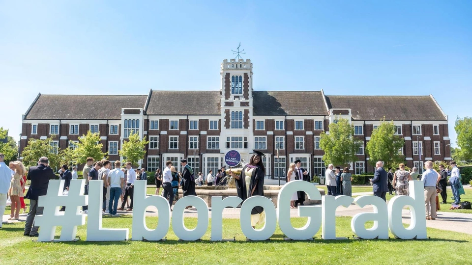 Photo of the Hazlerigg Building and lawn on a sunny day with big white letter signage spelling out ''#LboroGrad' on the lawn, with people behind it wearing graduation gowns
